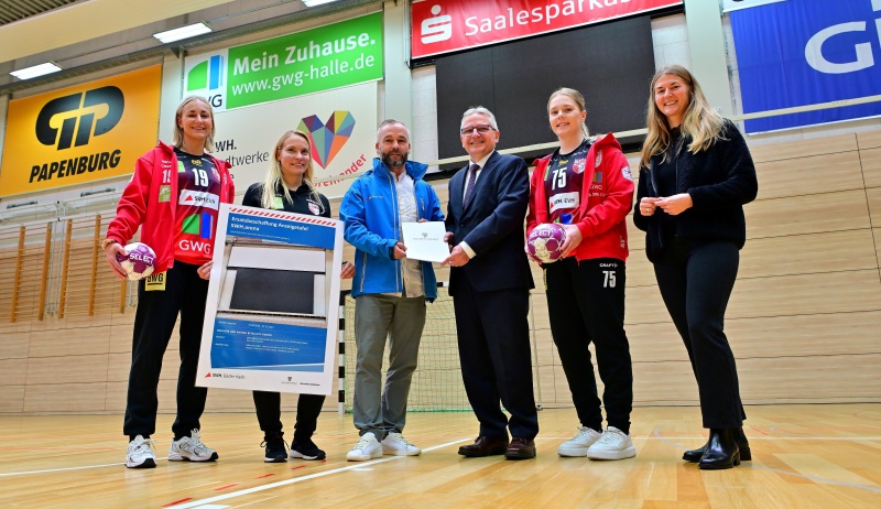 Lea Gruber (WILDCATS-Spielerin), Stefanie Kunze (Geschäftsstellenleiterin SV UNION Halle-Neustadt e.V. – WILDCATS), Nico Kanitz (Betriebsleiter Bäder Halle GmbH), Klaus Zimmermann (Staatssekretär Ministerium für Inneres und Sport Land Sachsen-Anhalt), Vanessa Dierks (WILDCATS-Spielerin) und Kerstin Godenrath (Mitglied des Landtages von Sachsen-Anhalt)stehen von links nach rechts nebeneinander in der SWH.arena und halten den Fördermittelbescheid, sowie zwei Handbälle in den Händen.