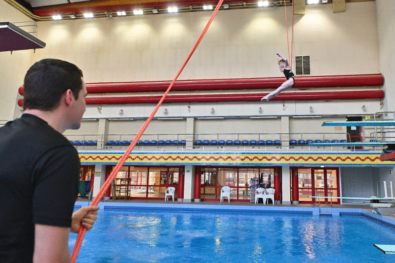 Ein Mann hält ein langes Seil über ein Schwimmbecken, an dem ein Mädchen in Turnkleidung hoch über dem Wasser schwingt.
