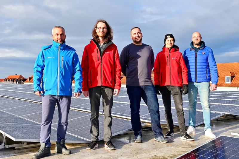 Fünf Männer stehen im Sonnenschein und mit dicken Jacken auf einem Dach. Auf dem Dach sind Solarplatten zu sehen. Alle blicken in die Kamera. Der Himmel ist leicht bewölkt.