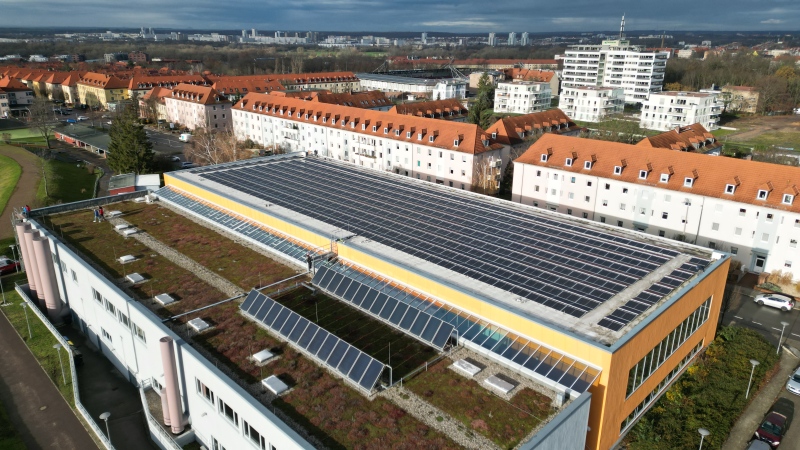 Eine Fotoaufnahme aus der Luft auf das Dach der Robert-Koch-Schwimmhalle. Außerdem ist dier Rest der stadt in der Vogelperspektive zu sehen. Der Himmel ist leicht bewölkt, aber die Sonne strahlt.