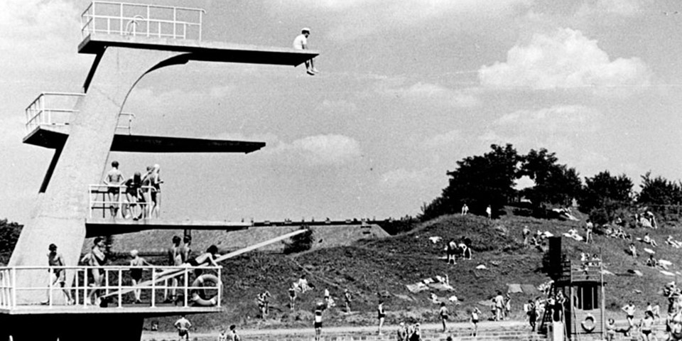 Eine schwarz-weiß-Aufnahme des Nordbades. Links ist der Sprungturm des Bades: auf der obersten Plattform sitzt eine Person und schaut nach unten. Auf den Plattformen zwei und drei stehen Menschen. Rechts im Bild ist das Becken mit Menschen, dahinter eine bergige Liegewiese mit Menschen. Am Himmel sind Wolken zu erkennen.