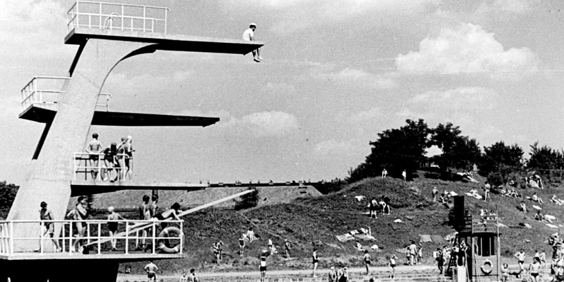 Eine schwarz-weiß-Aufnahme des Nordbades. Links ist der Sprungturm des Bades: auf der obersten Plattform sitzt eine Person und schaut nach unten. Auf den Plattformen zwei und drei stehen Menschen. Rechts im Bild ist das Becken mit Menschen, dahinter eine bergige Liegewiese mit Menschen. Am Himmel sind Wolken zu erkennen.