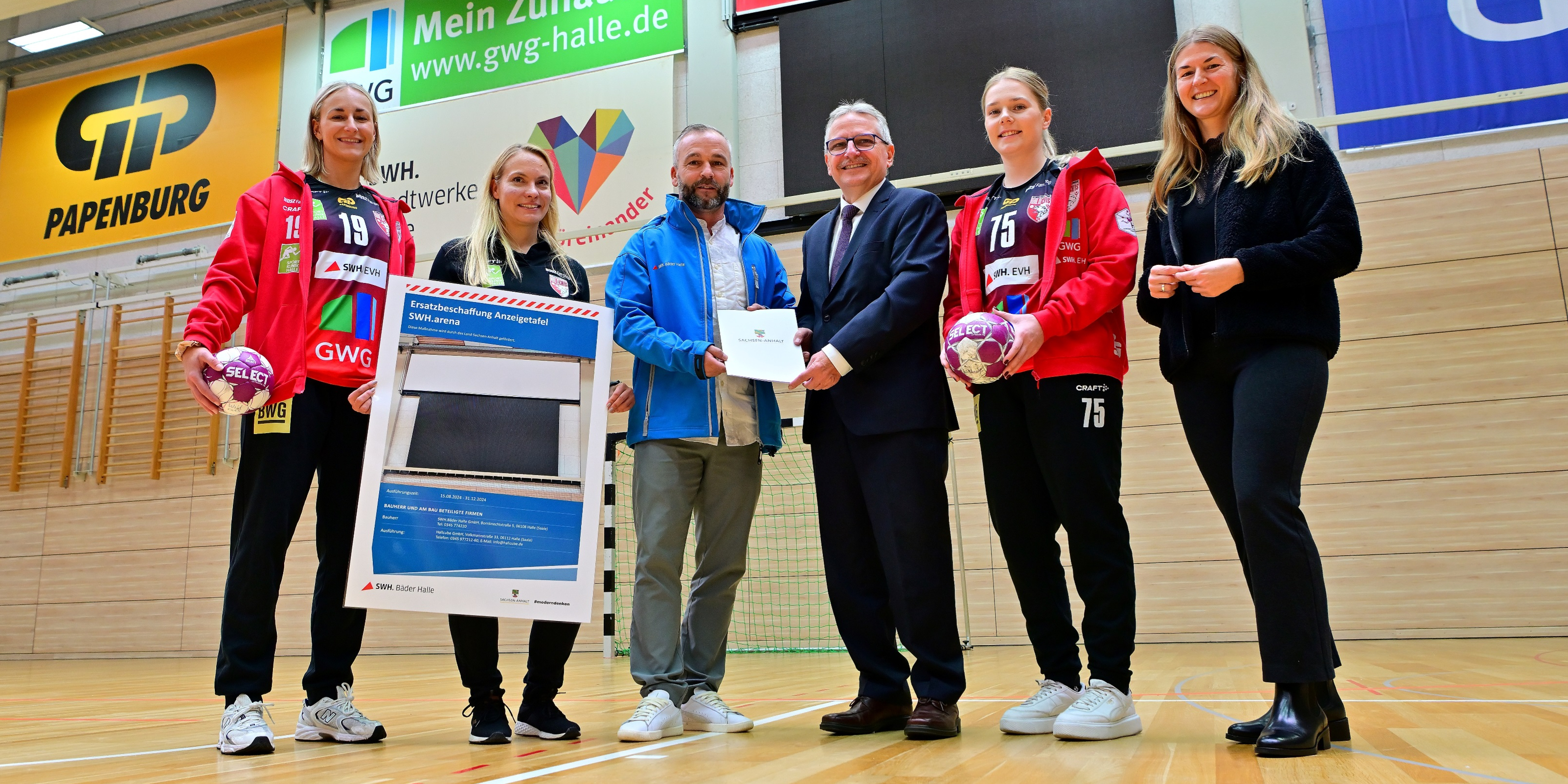 Lea Gruber (WILDCATS-Spielerin), Stefanie Kunze (Geschäftsstellenleiterin SV UNION Halle-Neustadt e.V. – WILDCATS), Nico Kanitz (Betriebsleiter Bäder Halle GmbH), Klaus Zimmermann (Staatssekretär Ministerium für Inneres und Sport Land Sachsen-Anhalt), Vanessa Dierks (WILDCATS-Spielerin) und Kerstin Godenrath (Mitglied des Landtages von Sachsen-Anhalt) stehen in einer Turnhalle und posieren mit dem Fördermittelbescheid.