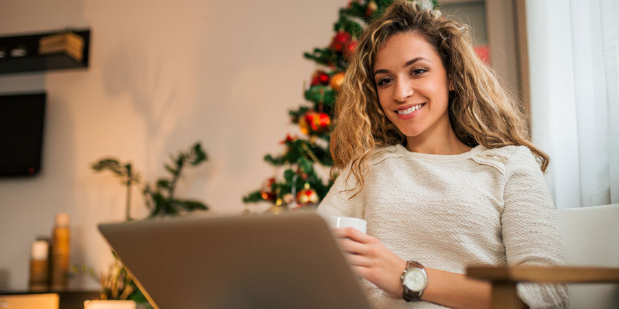 Eine junge Frau mit blondem lockigen Haar blick auf Ihren Laptop, der auf ihrem Schoß  steht. In der linken Hand hält Sie eine Tasse, sie lächelt. Im Hintergrund ist ihr Zimmer zusehen, in dem auch ein Weihnachtsbaum steht.