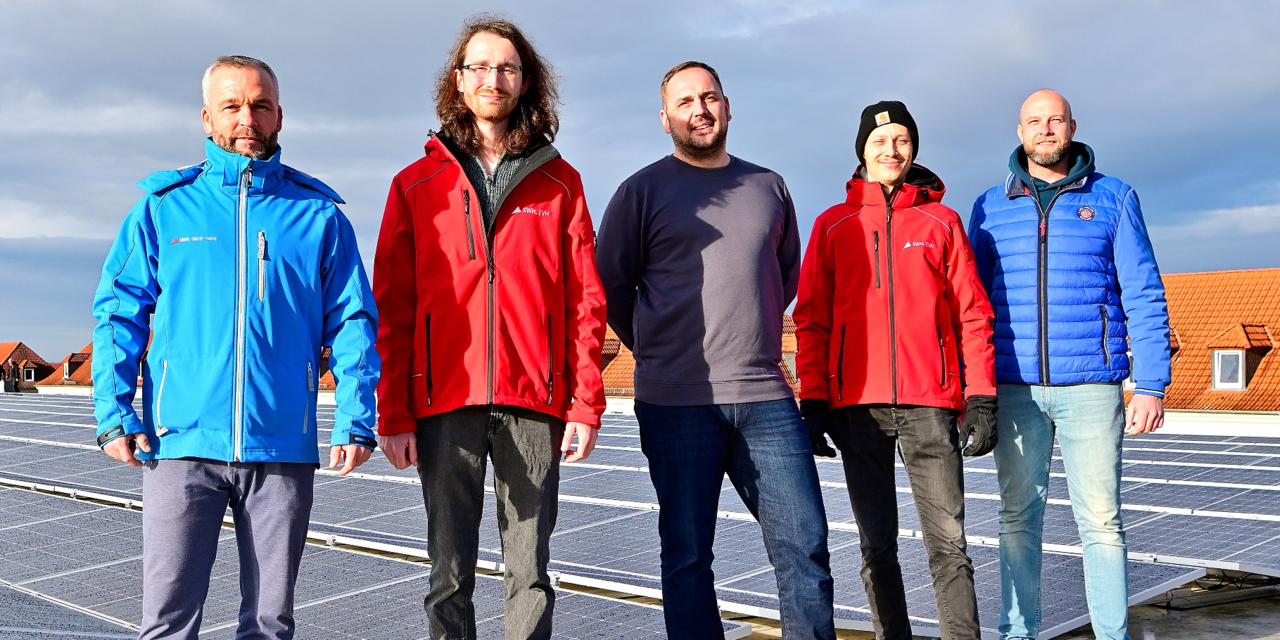 
Fünf Männer stehen im Sonnenschein und mit dicken Jacken auf einem Dach. Auf dem Dach sind Solarplatten zu sehen. Alle blicken in die Kamera. Der Himmel ist leicht bewölkt.
