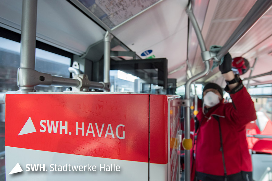 Ein Mitarbeiter mit roter Arbeitskleidung reinigt einen Teil eines Busses. Die Person ist verschwommen zu sehen. Im Vordergrund ist das Logo der HAVAG auf einem Fahrkartenautomaten zu sehen.