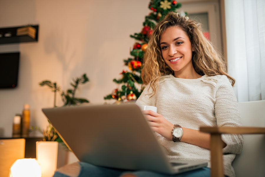 Eine junge Frau mit blondem lockigen Haar blick auf Ihren Laptop, der auf ihrem Schoß  steht. In der linken Hand hält Sie eine Tasse, sie lächelt. Im Hintergrund ist ihr Zimmer zusehen, in dem auch ein Weihnachtsbaum steht.