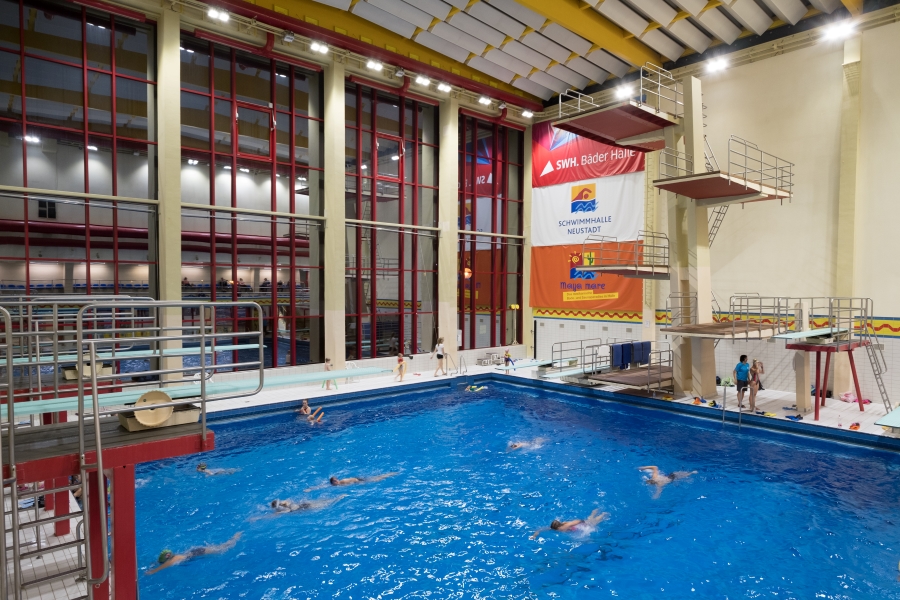 Ein Blick in die Neustädter Schwimmhalle: Die Fotoaufnahme wurde abends gemacht. Durch die große Fensterscheibe sieht es aus, als wäre es draußen schwarz. Am unten Bildrand ist das Wasser in dem Menschen schwimmen. Am rechten Bildrand sind die vier Absprung-Plattformen zu sehen. 