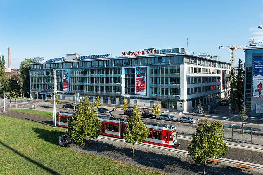 Eine Luftaufnahme des SWH-Gebäudes an der Spitze. Der Himmel ist blau. Im Vordergrund fährt eine rote Bahn der HAVAG. Am Gebäude hängen 2 Werbeplakate. Auf dem Dach sind Leuchtbuchstaben für den Unternehmensnamen "Stadtwerke Halle".