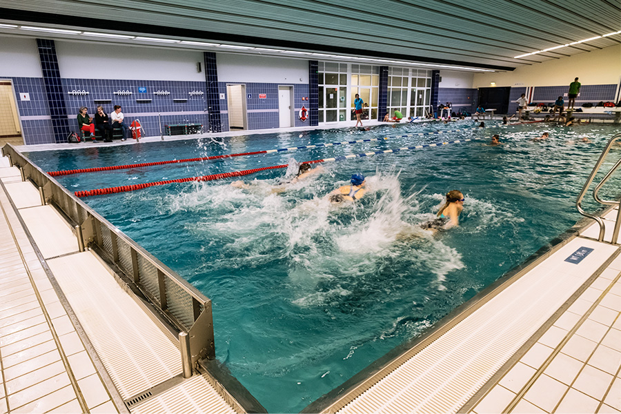 Kinder mit Schwimmbrille und -haube schwimmen nebeneinander im Wasser des Saline Hallenbades. Das Wasser spritzt. Im Hintergrund sind verschiedene Personen zu sehen: Badeaufsicht, und weitere Kinder. 