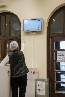 Eine Person zeigt auf einen an der Wand hängenden Bildschirm mit interaktiver Stadtkarte und Echtzeit-Daten zu Straßenbahnen und Bussen.