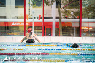 Schwimmerin im Becken mit Blick ins Wasser, im Hintergrund fährt vor der Schwimmhalle Neustadt eine Straßenbahn