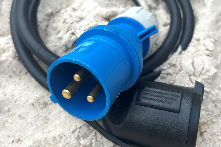 Blauer CEE-Stecker mit drei runden Metallstiften und schwarzem Kabel auf sandigem Untergrund