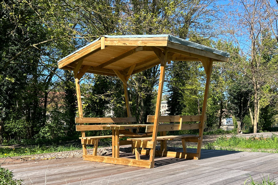 Holzüberdachung mit Sitzbänken und Tisch auf Holzterrasse vor Bäumen