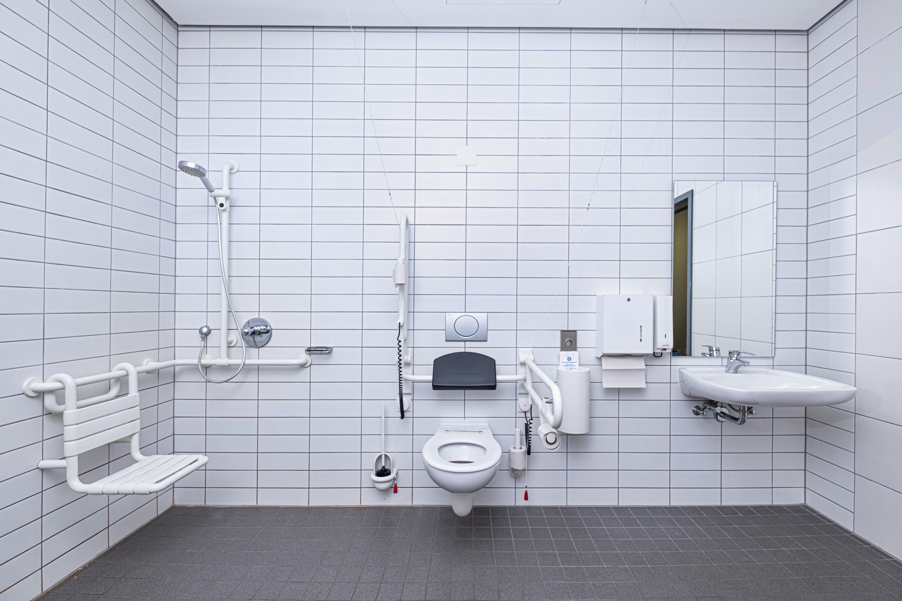 Das Bild zeigt den barrierefreien Sanitärbereich in der Robert Koch Schwimmhalle. Zu sehen ist eine ebenerdige Dusche mit Haltegriffen und Sitzmöglichkeit sowie eine Toilette mit Haltegriffen und Abstützhilfen.