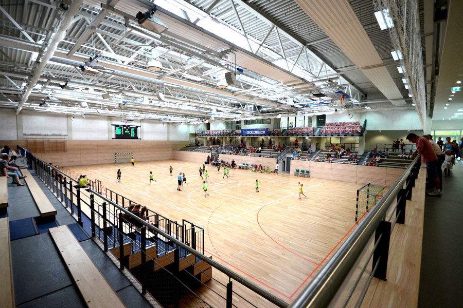 Innenansicht einer großen Sporthalle mit Holzboden, Tribünen und mehreren spielenden Personen auf dem Spielfeld