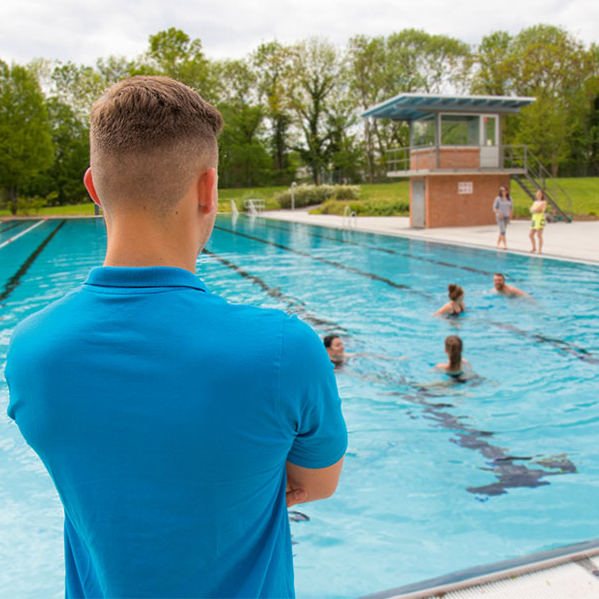 Ein Mitarbeiter steht mit dem Rücken zur Kamera und blickt über das Schwimmbecken. Im Wasser schwimmen mehrere Personen.