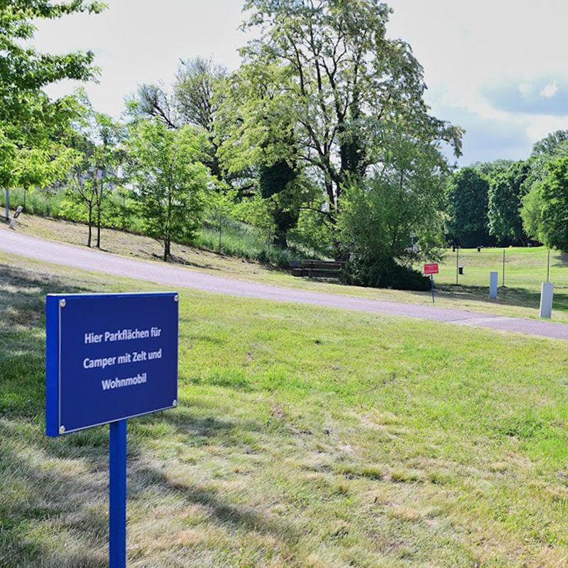 Grüne Wiese mit mehreren Schildern, eines im Vordergrund mit weißer Schrift auf blauem Grund, Bäume und ein Weg im Hintergrund