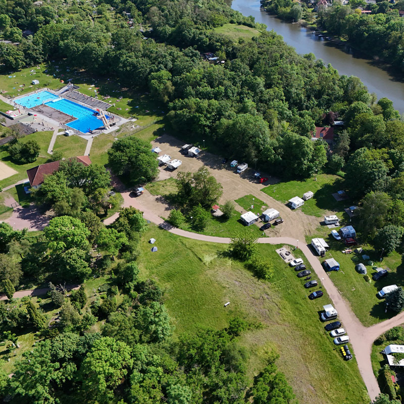 Luftaufnahme eines Campingplatzes mit Wohnmobilen und Zelten an einem Flussufer neben einem Freibad