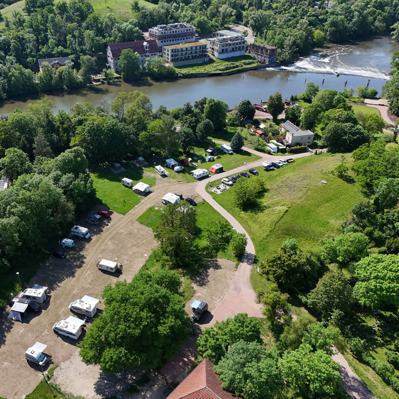 Luftaufnahme eines Campingplatzes mit Wohnmobilen und Zelten an einem Flussufer mit kleinem Wehr