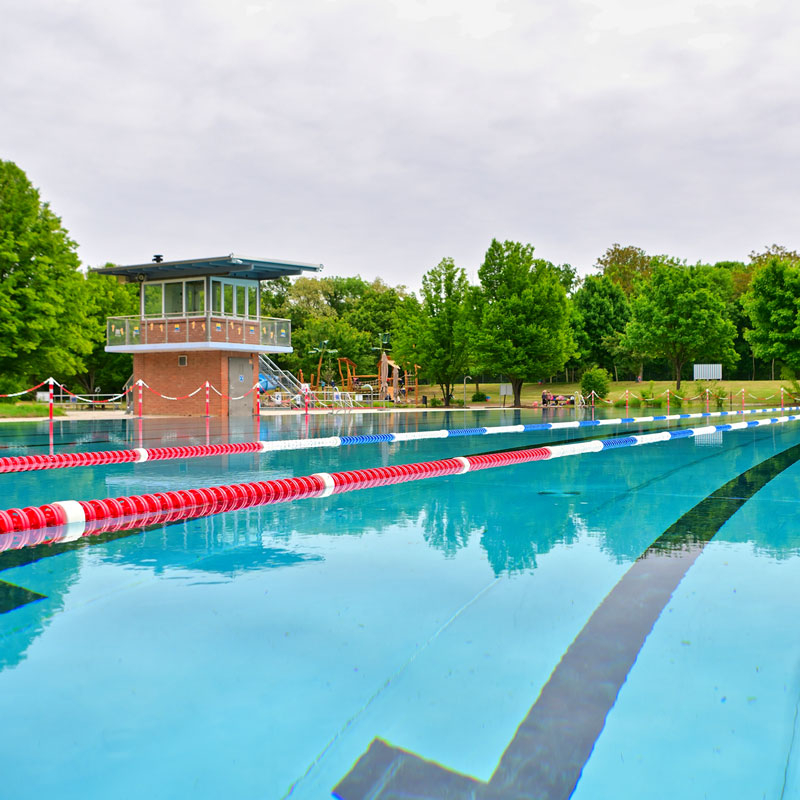 Freibad mit Schwimmbecken, Retttungsschwimmerturm und Liegewiese mit Bäumen im Hintergrund
