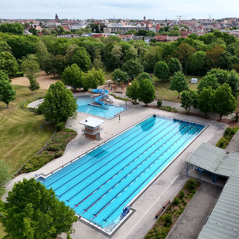 Luftaufnahme des Freibads mit großem Schwimmbecken, kleinem Becken mit Wasserrutsche und umgebenden Bäumen vor einer Stadtkulisse