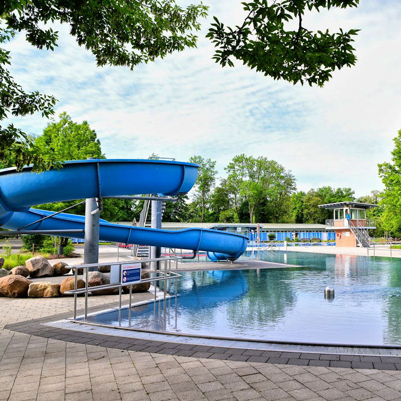 Freibad mit blauer Wasserrutsche, Schwimmbecken und Umkleidegebäude im Hintergrund unter bewölktem Himmel