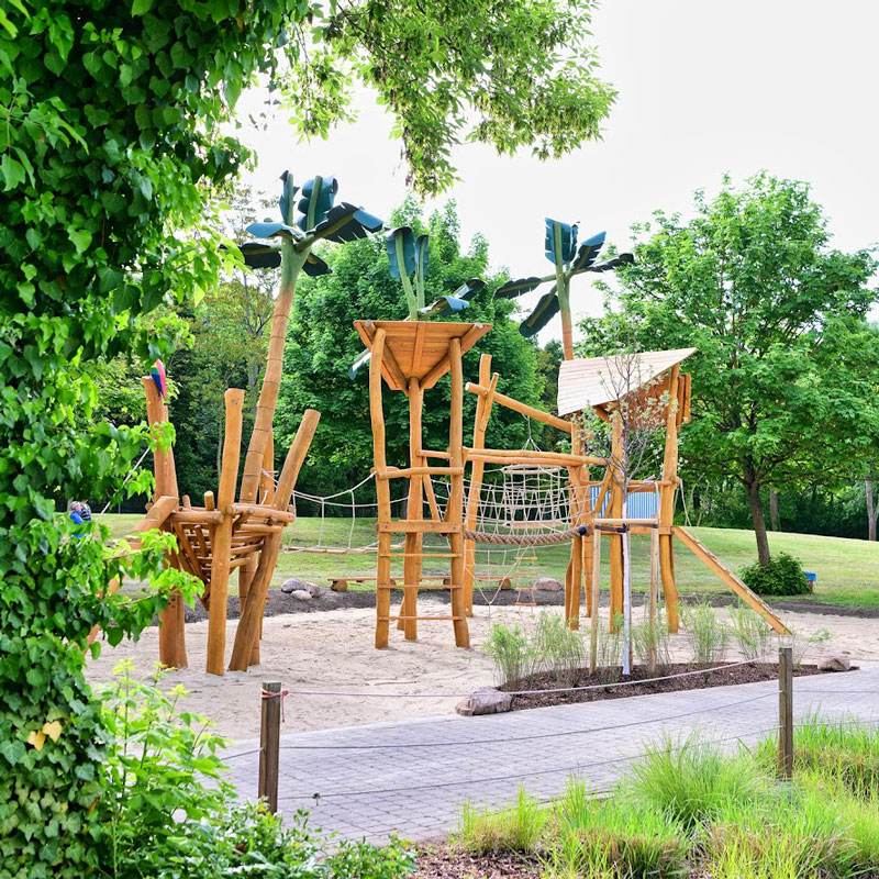 Spielplatz mit Klettergerüst aus Holz und Netz im Sandbereich umgeben von Bäumen und Grünflächen
