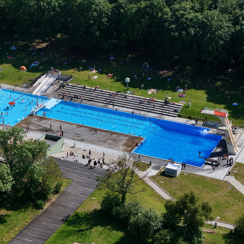Luftaufnahme eines Freibads mit mehreren Schwimmbecken, Sprungturm und umliegenden Liegewiesen in einem grünen Park