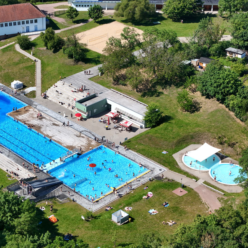 Luftaufnahme eines Freibads mit mehreren Schwimmbecken, Sprungturm und umliegenden Liegewiesen in einem grünen Park