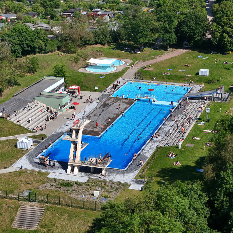Luftaufnahme eines Freibads mit mehreren Schwimmbecken, Sprungturm und umliegenden Liegewiesen in einem grünen Park