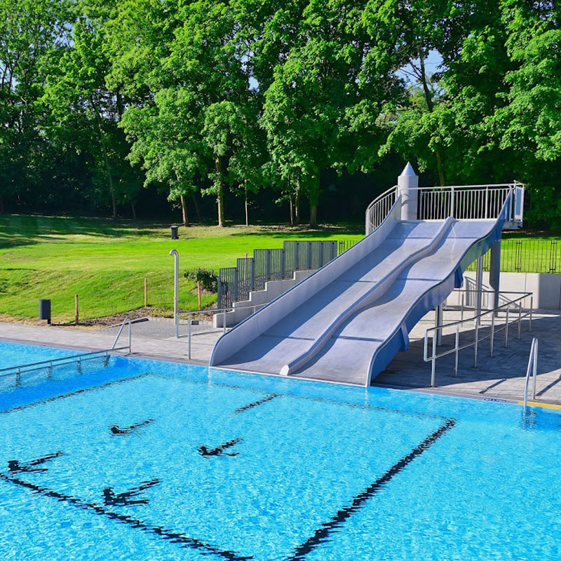 Zweispurige Wasserrutsche mit Treppe und Geländer neben einem Schwimmbecken im Freibad mit Bäumen im Hintergrund