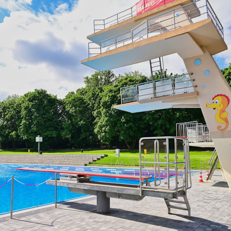 Sprungturm mit mehreren Plattformen und einem Sprungbrett an einem Freibad, auf der Seite ein Seepferdchen-Motiv, im Hintergrund Bäume und blauer Himmel mit Wolken