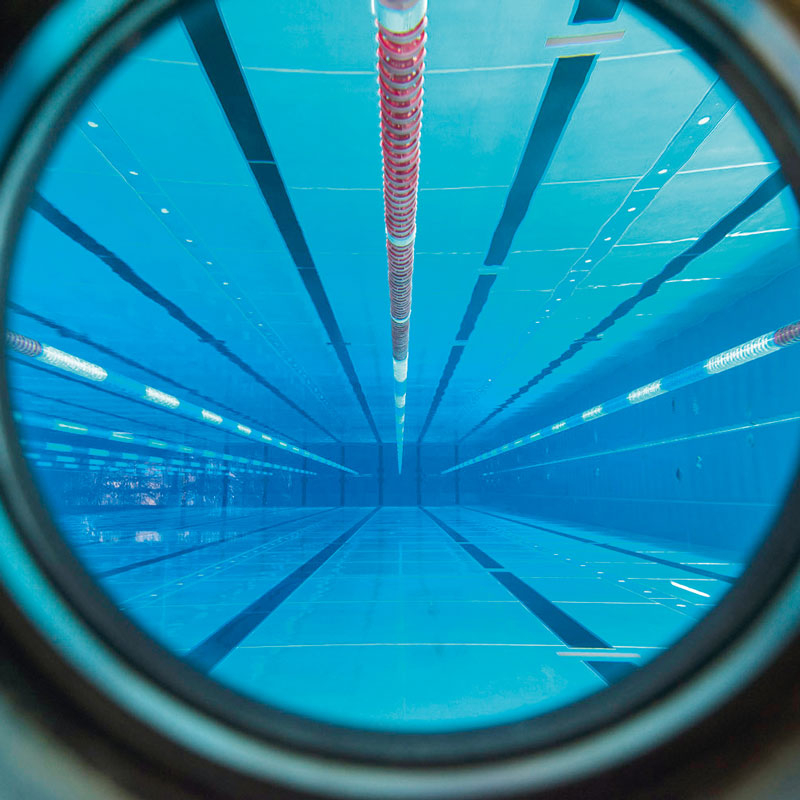 Blick durch ein rundes Sichtfenster auf ein leeres Schwimmbecken mit Bahnmarkierungen und einer roten Bahntrennleine in der Mitte