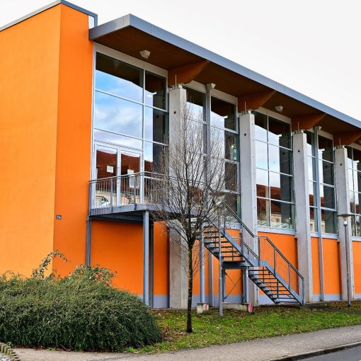 Außenansicht eines modernen Gebäudes mit oranger Fassade, großen Fenstern und einer Außentreppe neben einem Baum