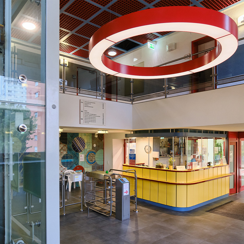 Empfangsbereich der Schwimmhalle Neustadt mit gelber Rezeption, Drehkreuz, Wegweiser und markanter roter Deckenleuchte.

