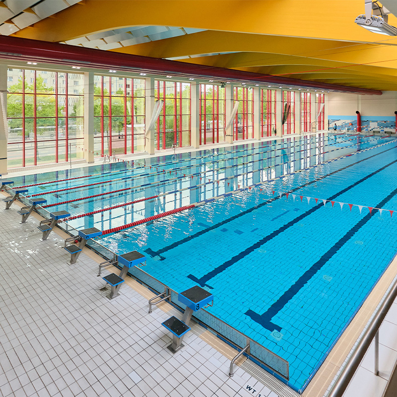 Innenansicht der Schwimmhalle Neustadt mit mehreren durch Schwimmleinen abgetrennten Bahnen, Startblöcken und großen Fenstern entlang des Beckens.