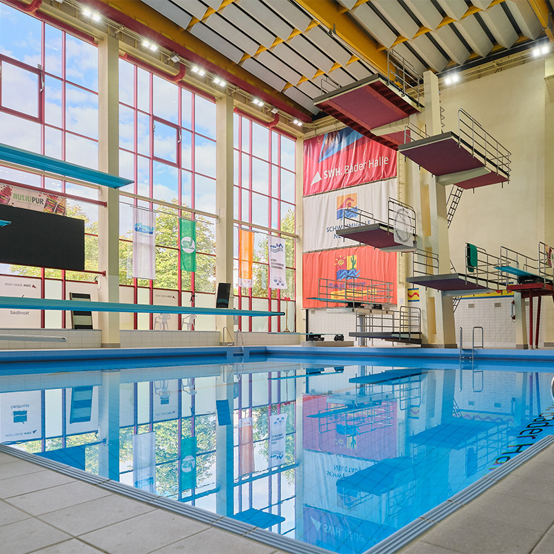 Innenansicht der Schwimmhalle Neustadt mit Sprungbecken, mehreren Sprungbrettern und Sprungplattformen sowie großen Fenstern.