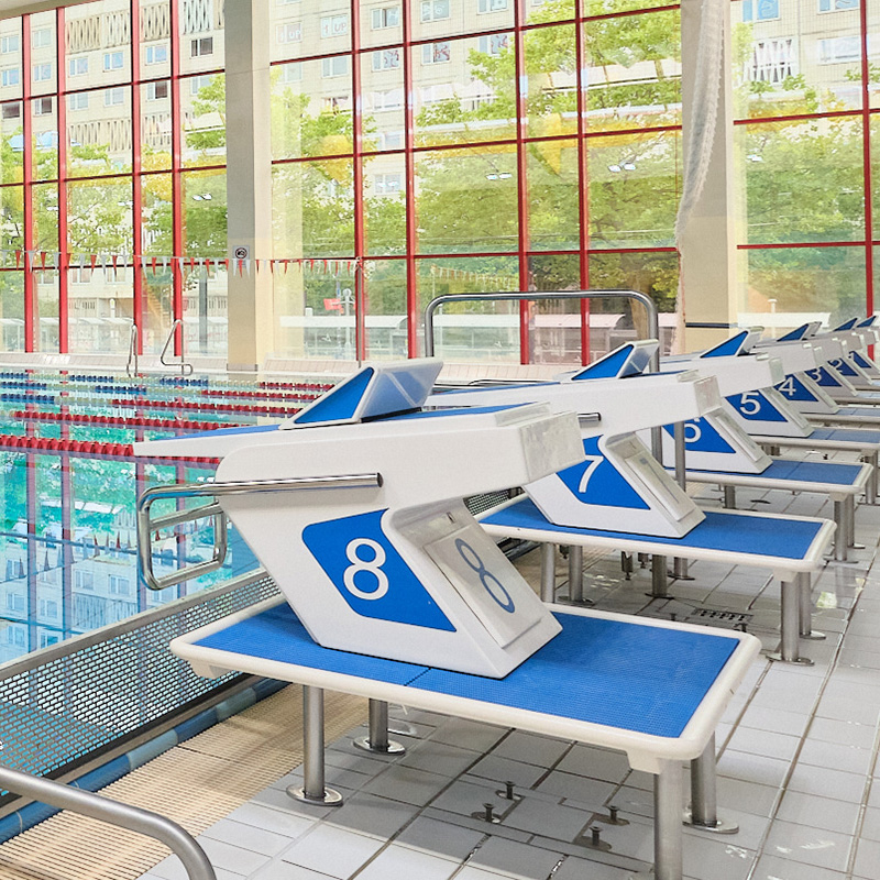 Reihe nummerierter Startblöcke am Schwimmbecken der Schwimmhalle Neustadt; im Hintergrund sind Schwimmbahnen, große Fenster und Wohngebäude zu sehen.