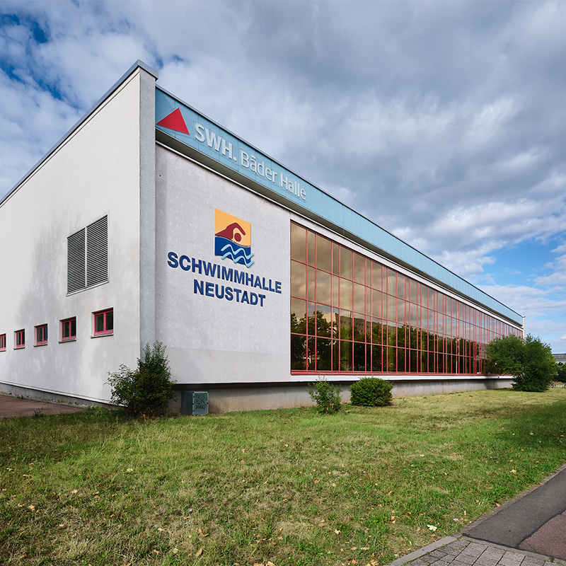 Außenansicht der Schwimmhalle Neustadt in Halle (Saale) mit großer Fensterfront, dem Schriftzug „Schwimmhalle Neustadt“ und dem Logo „SWH Bäder Halle“.