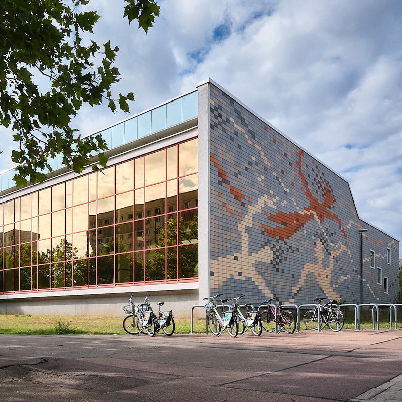 Außenansicht der Schwimmhalle Neustadt in Halle (Saale) mit großer Glasfassade, farbigem Fliesenmosaik an der Seitenwand und Fahrrädern vor dem Gebäude.