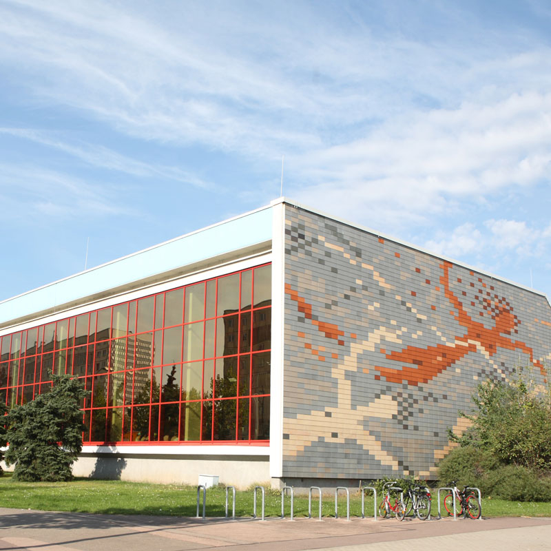 Außenansicht eines Gebäudes mit großen rot gerahmten Fenstern und einer Fassade aus grauen Kacheln mit orangefarbenem Muster