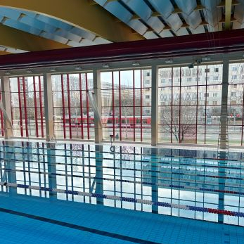 Ansicht eines Schwimmbeckens mit Bahnmarkierungen und großen Fenstern, durch die eine Stadtlandschaft sichtbar ist
