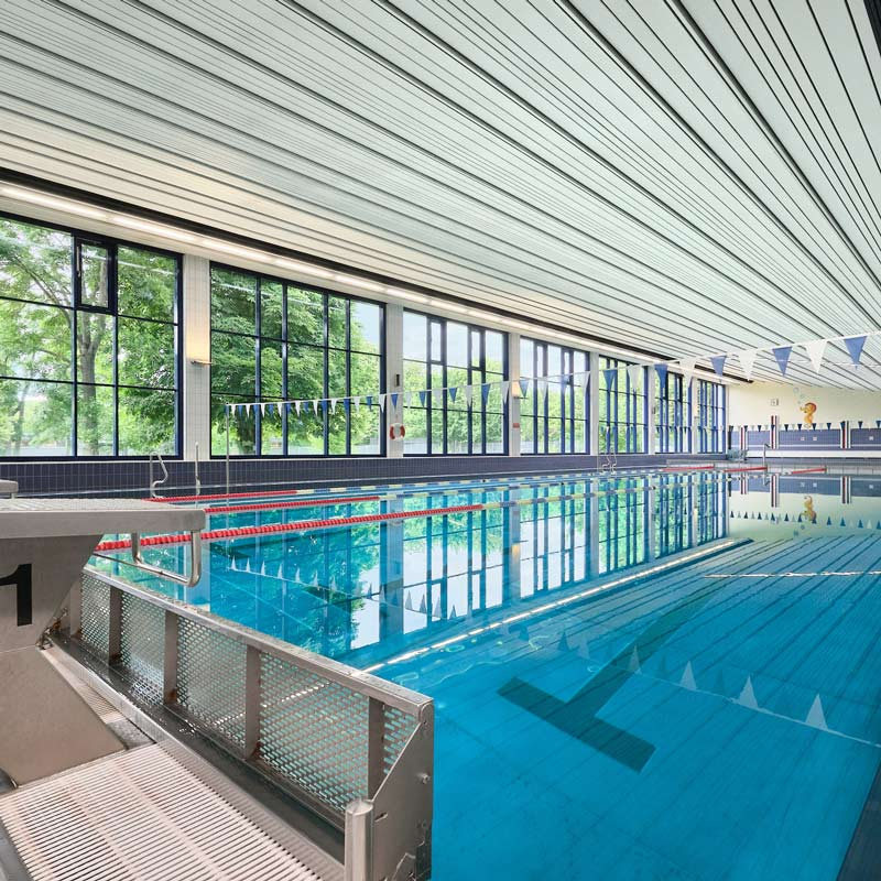 Innenansicht eines Schwimmbeckens mit Startblöcken, Bahnleinen und großen Fenstern, die Bäume draußen zeigen