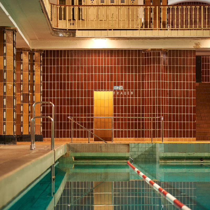 Ansicht des Hallenbades mit Wasserbecken, Treppengeländer und braun gefliester Wand mit Tür
