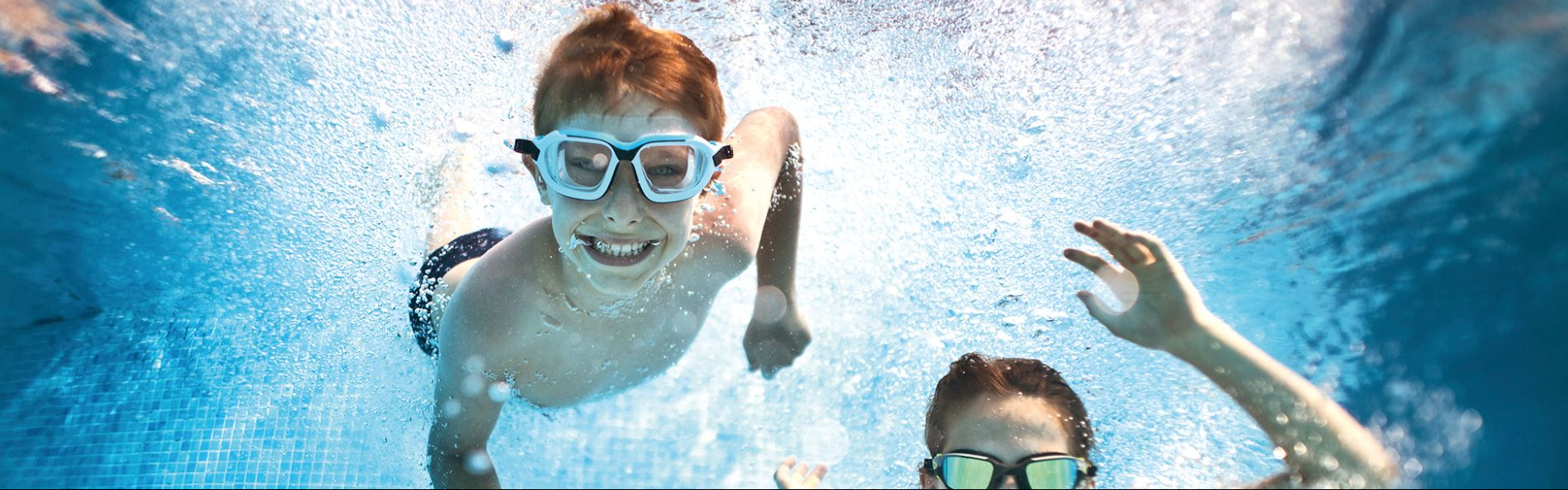 Zwei Kinder tauchen unter Wasser, die Kamera befindet sich auch unter Wasser. Die Kinder tragen Schwimmbrillen und lachen in die Kamera.